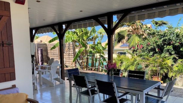 A covered outdoor dining area with a table and chairs, surrounded by lush greenery and a view of a residential area. - Villa Soleil Located in a quiet, serene residential area (Vauclin)