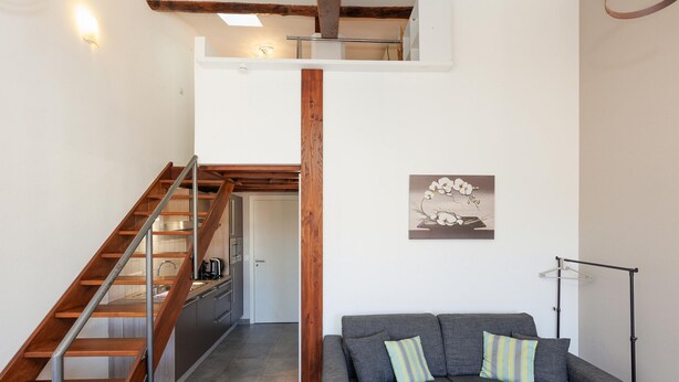 Ein modernes Interieur mit einer Holztreppe, einem grauen Sofa mit gestreiften Kissen und einer minimalistischen Einrichtung, darunter ein gerahmtes Kunstwerk an der Wand.
