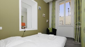 Una camera d'albergo con un letto grande, uno specchio, una finestra con tende e una piccola sedia con un cuscino.