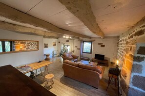 Ein rustikales Interieur mit sichtbaren Balken, einer Steinwand, einer Couch, einem Esstisch und einem Fernseher.