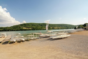 Ein Steg am Seeufer mit Booten, ein Sandstrand und ein bewaldeter Hügel im Hintergrund.