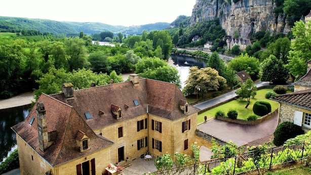 Un bâtiment historique en pierre, doté d’une cour intérieure, entouré d’une végétation luxuriante et bordé par une rivière.