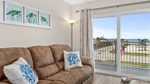 Een woonkamer met een bruine bank, twee decoratieve kussens, een lijstwerk met daarop schilderingen van zeeschildpadden, en uitzicht op het strand via een balkon met een hekwerk.