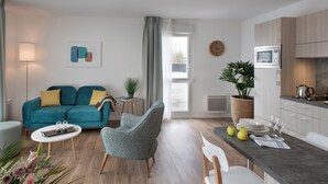 A modern living room with a teal sofa, a grey armchair, and a white coffee table. A kitchen area with a microwave and cabinets is visible to the right. - DOMITYS - La Flânerie (Béthune)
