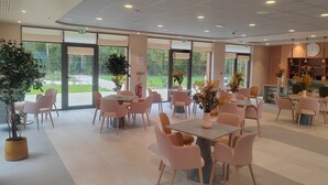 A spacious indoor area with multiple tables and chairs, large windows offering a view of the outdoors, and ceiling lights. - DOMITYS - La Flânerie (Béthune)