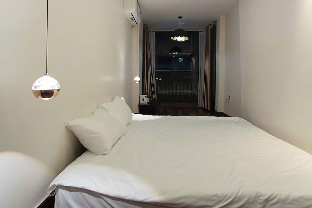 A modern hotel room with a large bed, a hanging light fixture, and a view of the dining area.
