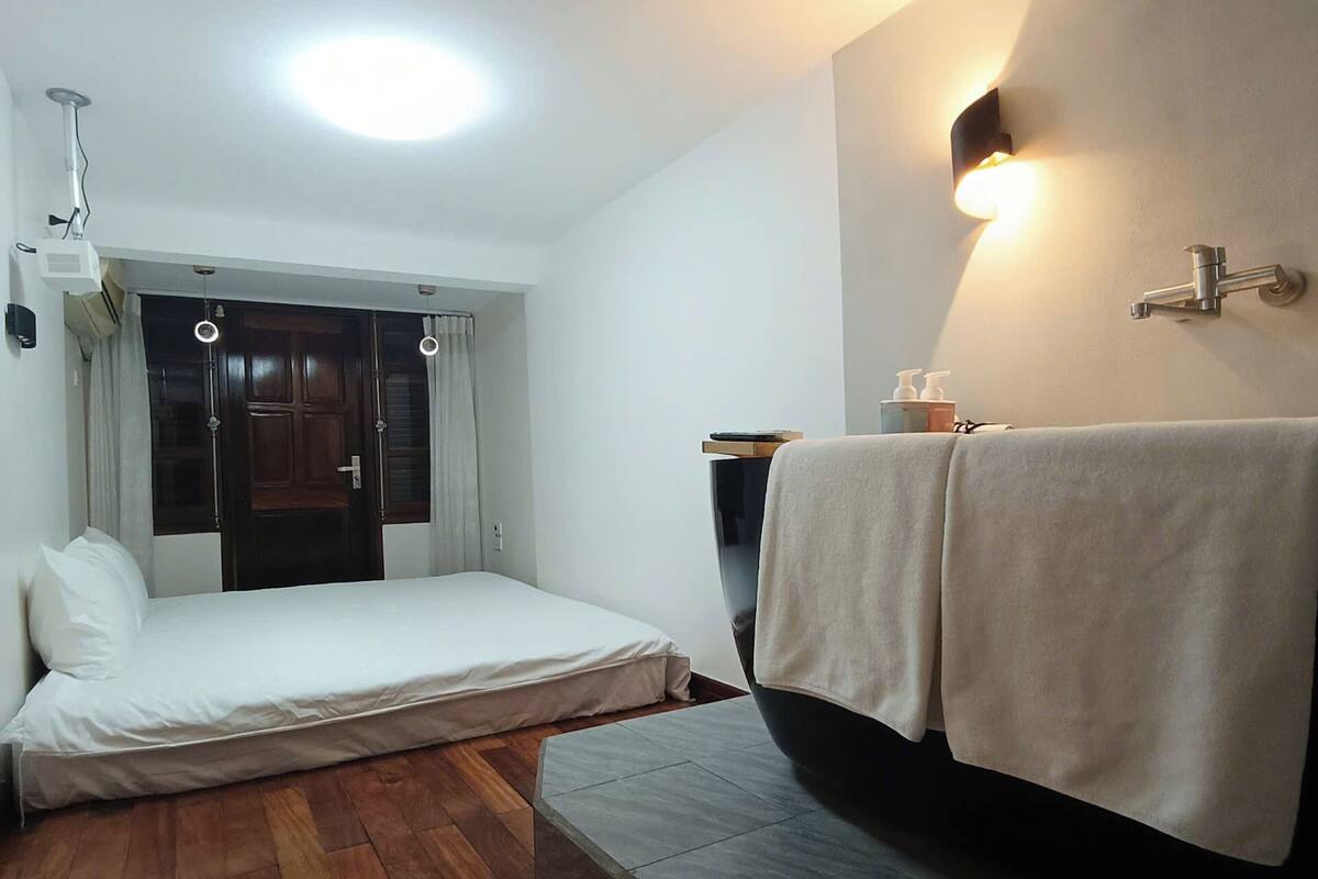 A compact room with a bed, a small table with toiletries, and a wall-mounted light fixture.