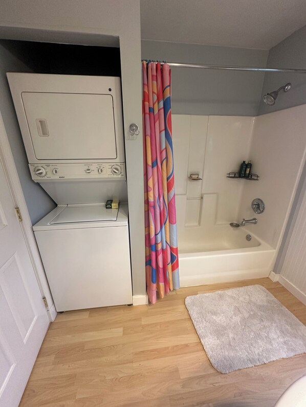 A compact bathroom with a built-in washer and dryer, a shower with a colorful curtain, and a small white rug on the floor.