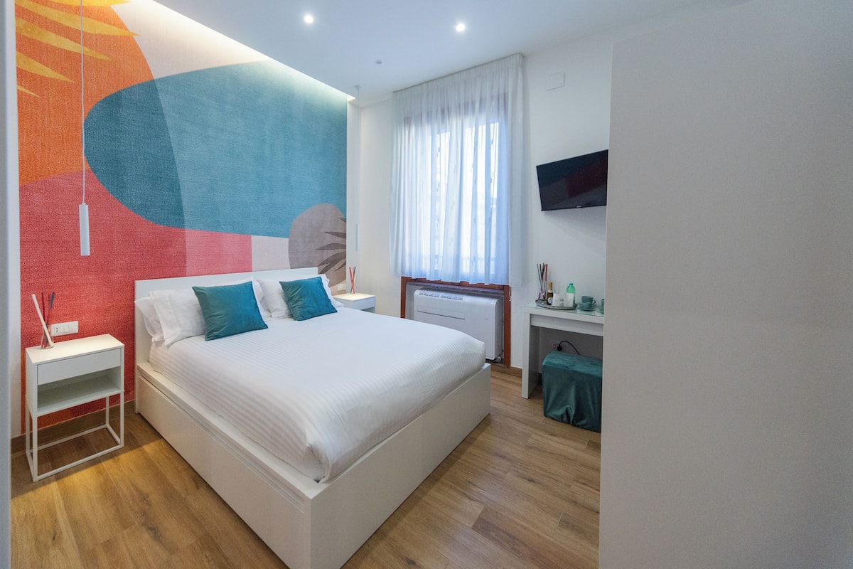 A modern hotel room with a large bed, a desk with a chair, a flat-screen TV, and a large abstract mural on the wall.