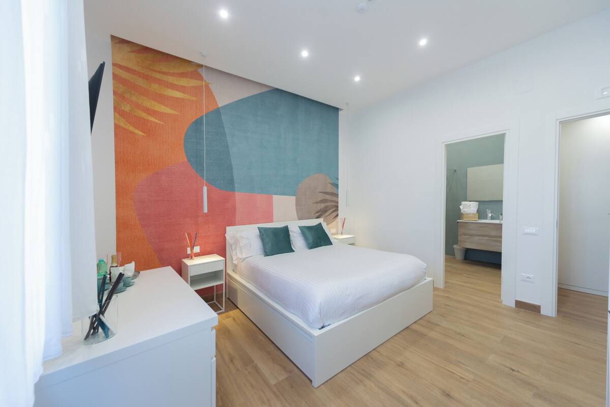 A modern bedroom with a large bed, a nightstand, a desk, and a wall mural.