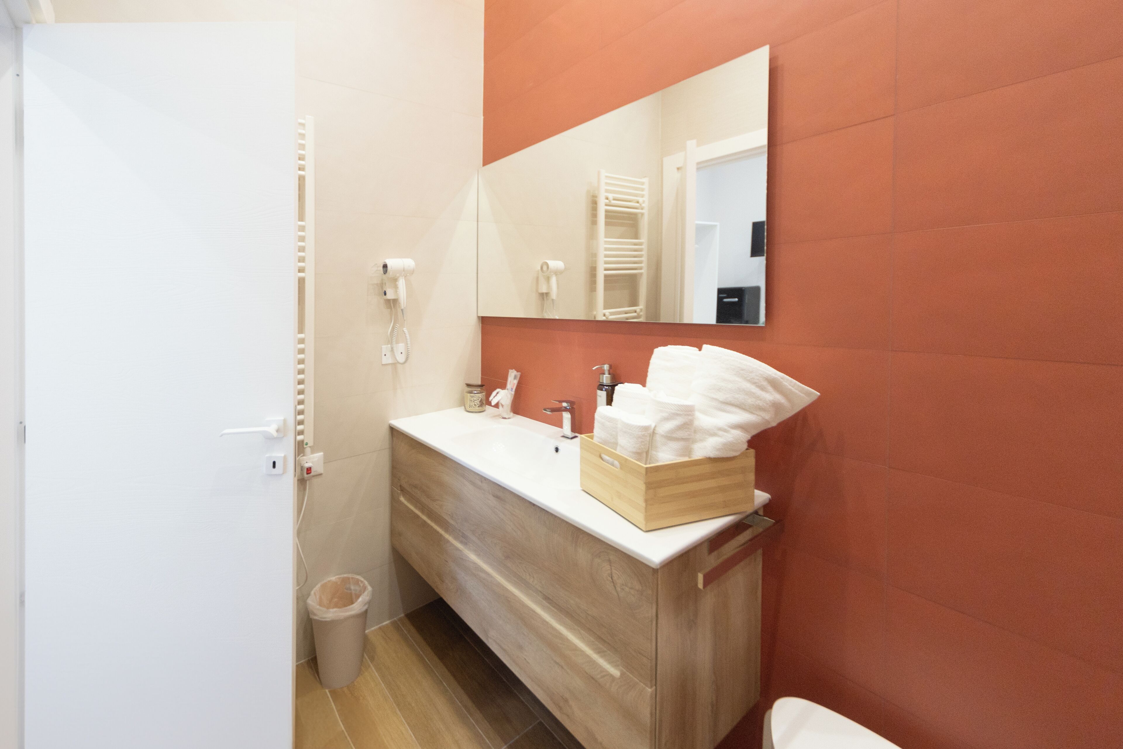 A bathroom with a wooden vanity, white sink, and a mirror. There is a trash can and a towel rack visible.