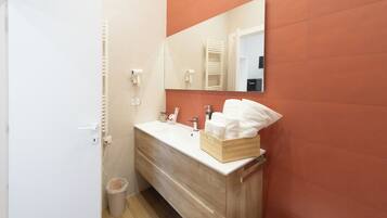 Une salle de bain avec un meuble-lavabo en bois, un lavabo blanc et un miroir. On peut voir une poubelle et un porte-serviettes.