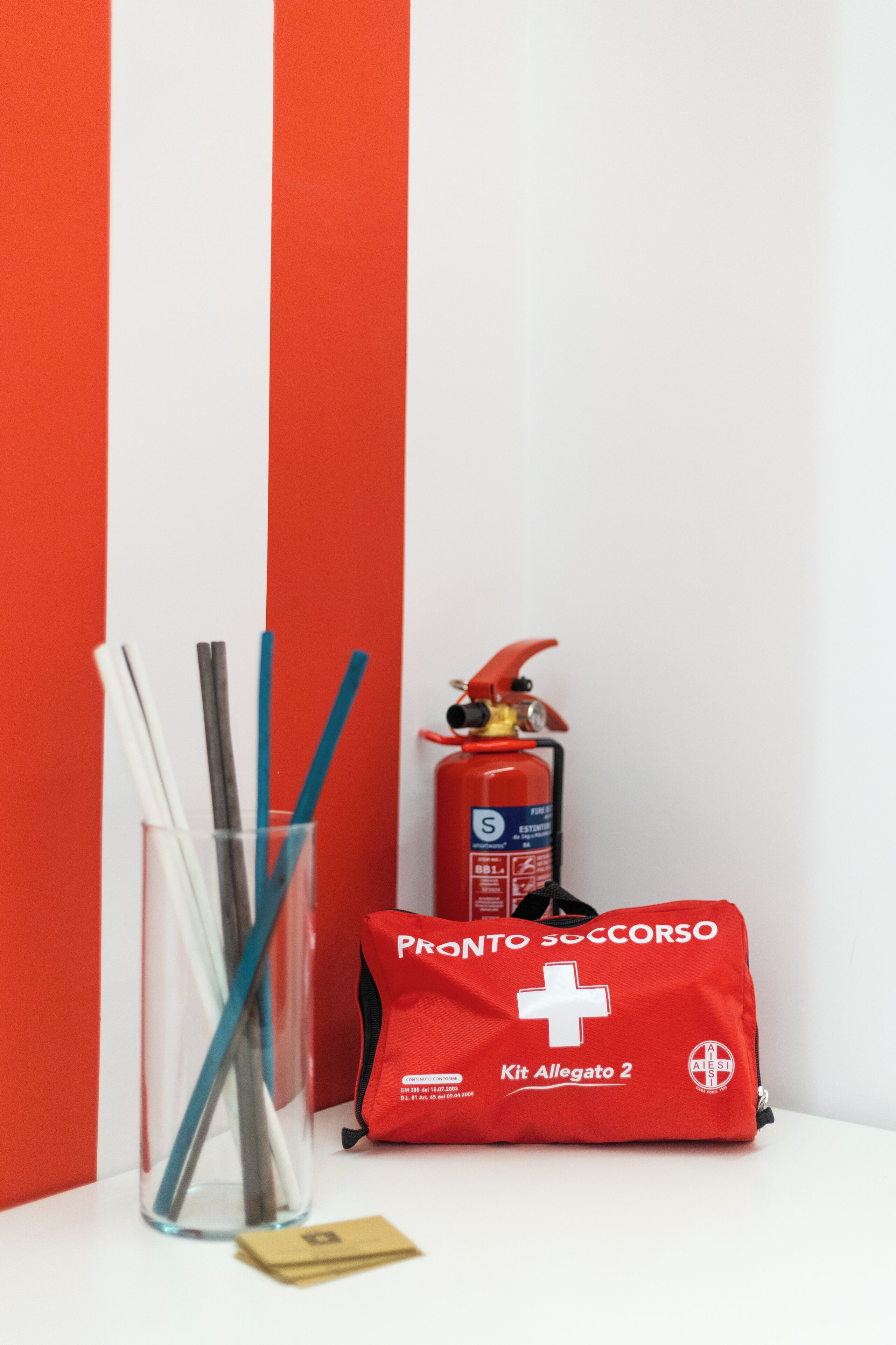 Une trousse de premiers secours rouge, un verre avec des pailles et un extincteur, le tout sur un fond rayé rouge et blanc.