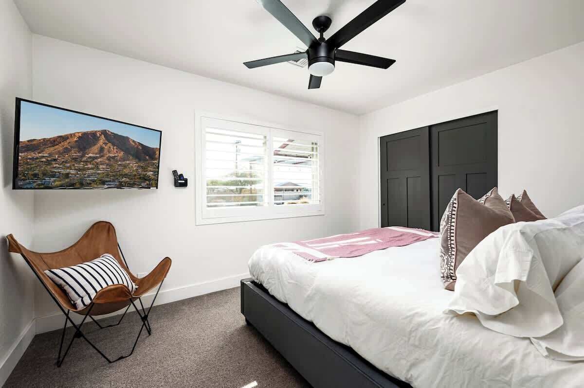 A bedroom with a bed, a ceiling fan, a window with blinds, a mounted TV, and a chair.