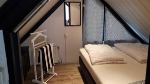 Quarto com teto inclinado, cama com roupa de cama branca, cabide com toalha listrada e janela com persianas verticais.