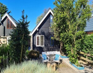 A two-story house with a dark exterior, white trim, and a small front porch with two white chairs and a blue cushion. - Holiday Home With Large Terrace (Sint Maartensvlotbrug)
