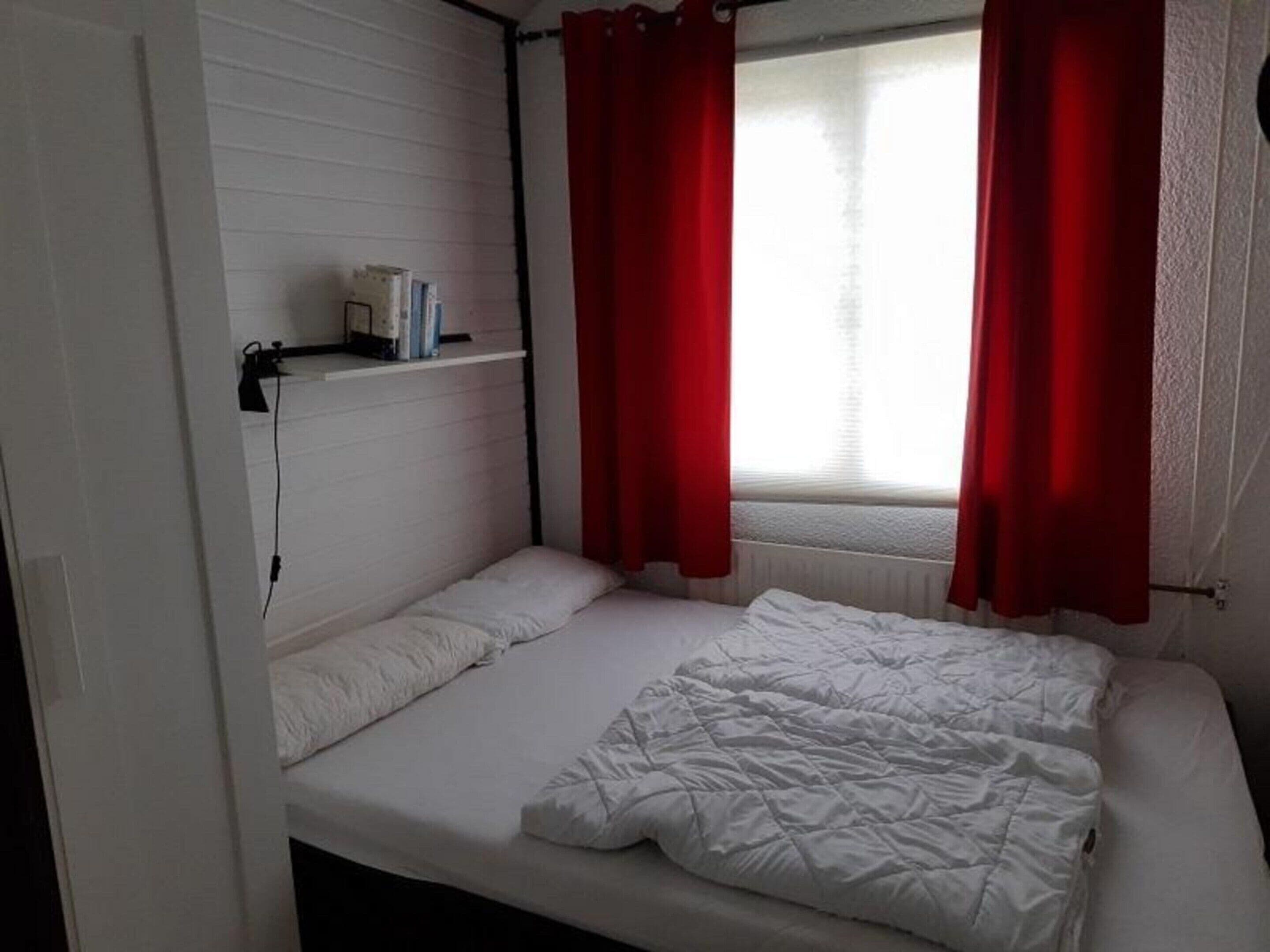 A small, single-bed room with white bedding, a shelf with books, and red curtains on a window.