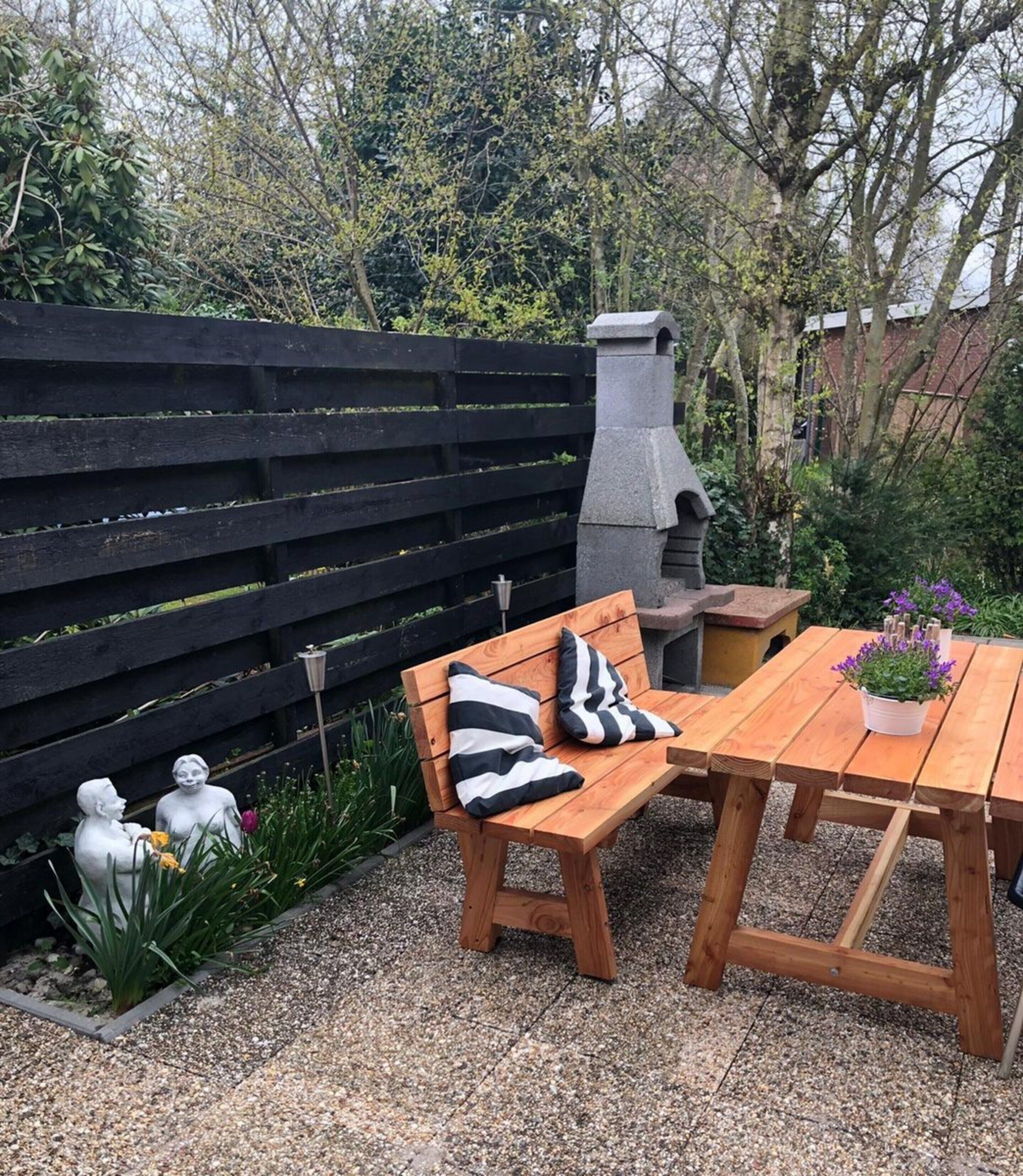 A wooden bench with striped cushions, a stone birdbath, and a table with a potted plant.