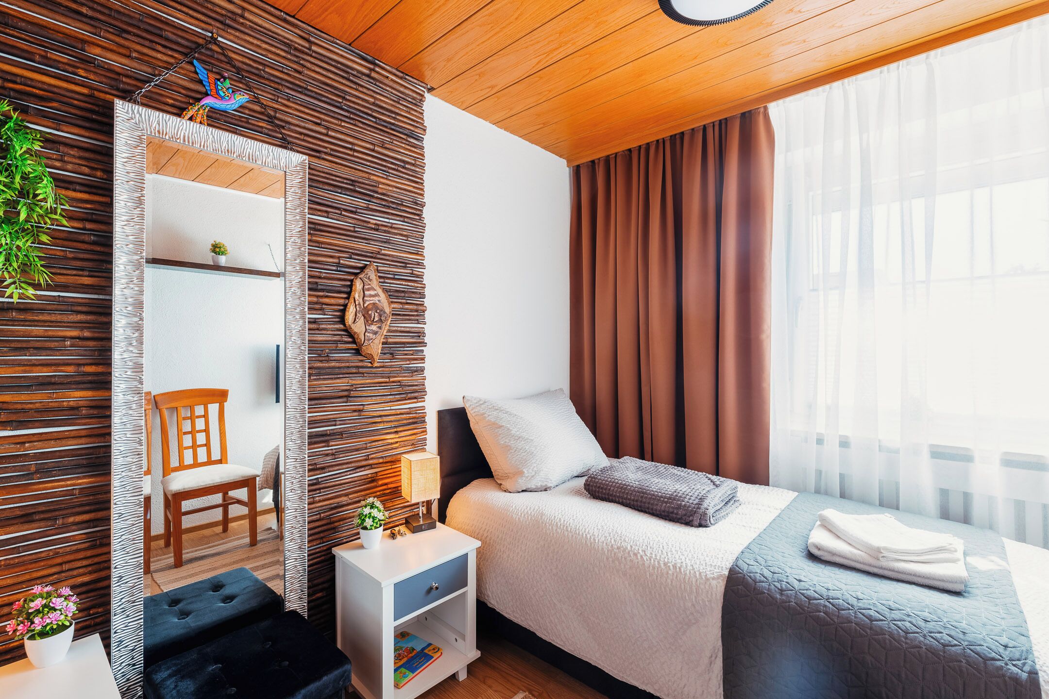 A modern bedroom with a wooden ceiling, a bed with a grey quilt, a nightstand with a lamp, a chair, and a mirror with a decorative frame.