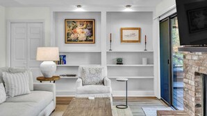 A living room with a sofa, armchair, coffee table, and fireplace. There are shelves with decorative items and a painting on the wall.