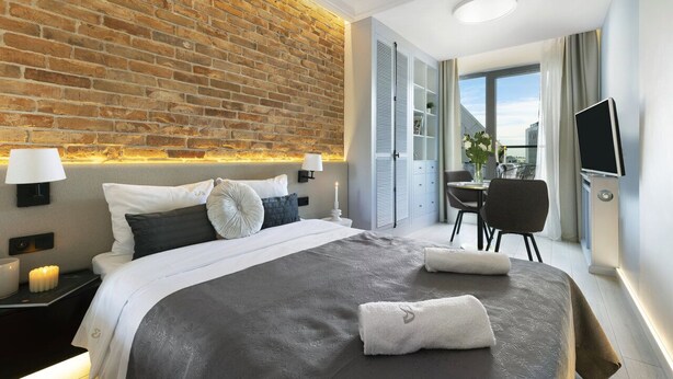 Ett modernt hotellrum med en tegelvägg, en säng med ett grått och vitt täcke, ett sängbord med en lampa, en tv och utsikt över staden genom stora fönster.