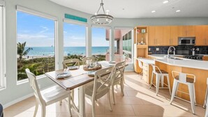 Une cuisine moderne avec de grandes fenêtres, un coin repas avec des chaises blanches et une vue sur la plage.