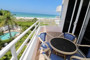 A balcony with a table and chairs, overlooking a beach and a pool.
