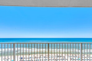 Vista de una playa desde un balcón con barandilla, cielo azul despejado y una playa de arena con gente y sombrillas.