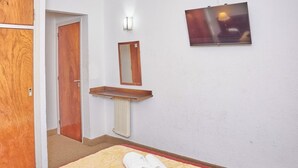 Une chambre d’hôtel avec une porte en bois, une petite étagère en bois, un miroir fixé au mur, un radiateur et un lit avec une serviette enroulée.