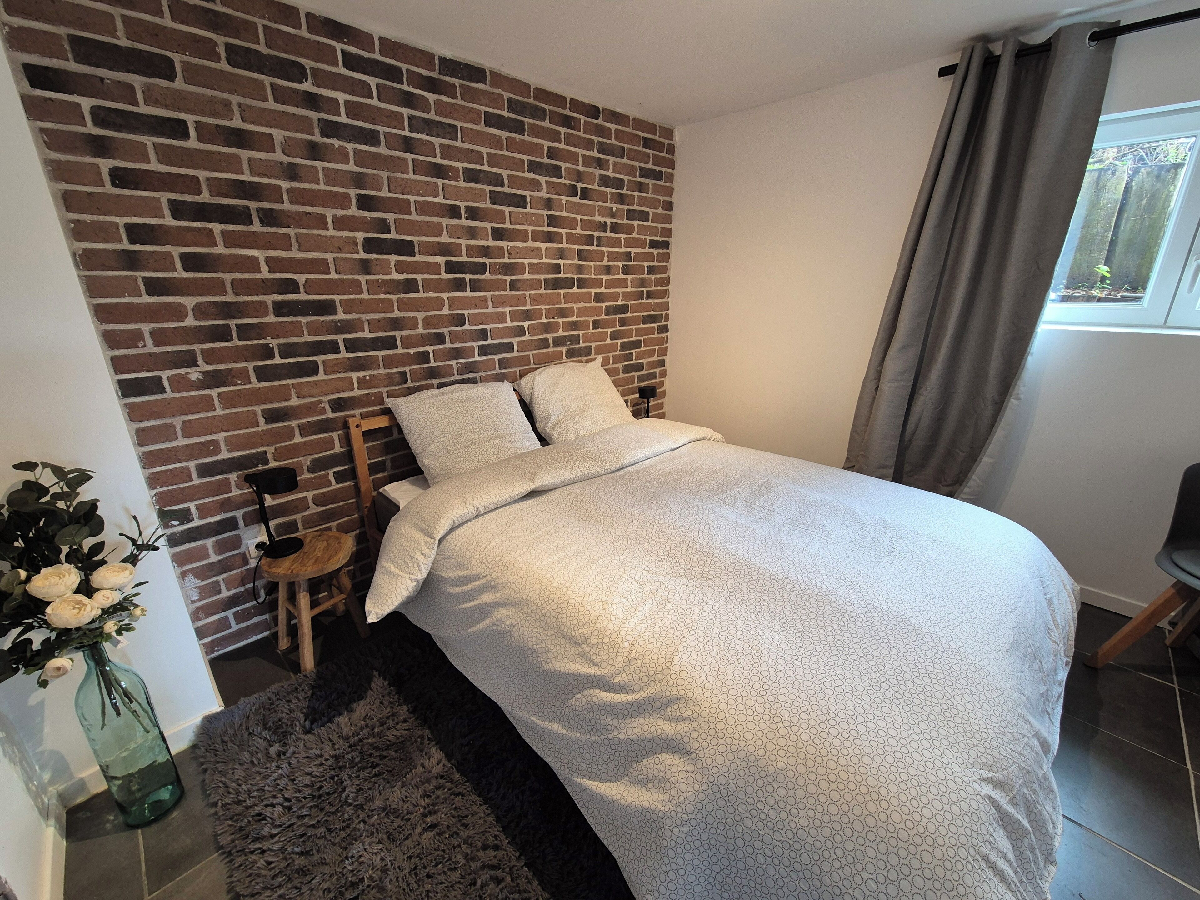 A bedroom with a brick-patterned wall, a bed with a white textured duvet, a small wooden nightstand, a vase with flowers, and a window with curtains.