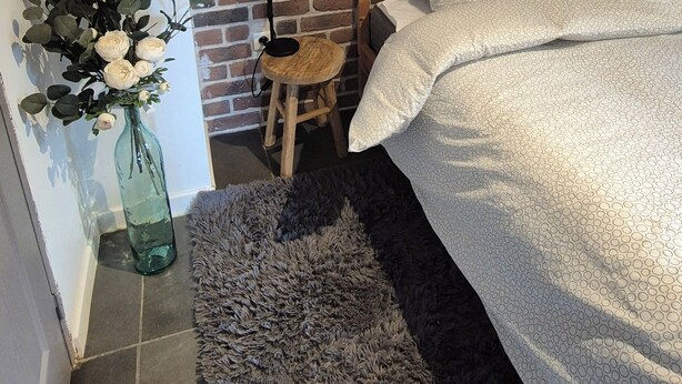 A bedroom with a brick wall, a wooden bed frame, a small stool, a green vase with flowers, and a fluffy rug.