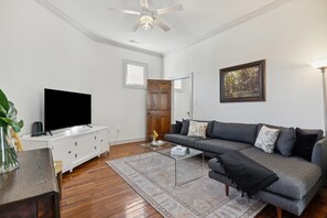 A living room with a flat-screen TV, a sofa, a coffee table, and a ceiling fan. - Pinkadilly on Spring! Walk to King St|Porch|Bikes (Charleston)