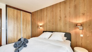 A bedroom with a wooden wall, a bed with white linens, two wall-mounted lamps, and a small round ottoman. - Apartment Mont d'Arbois Megeve - Ski in/out (Megève)