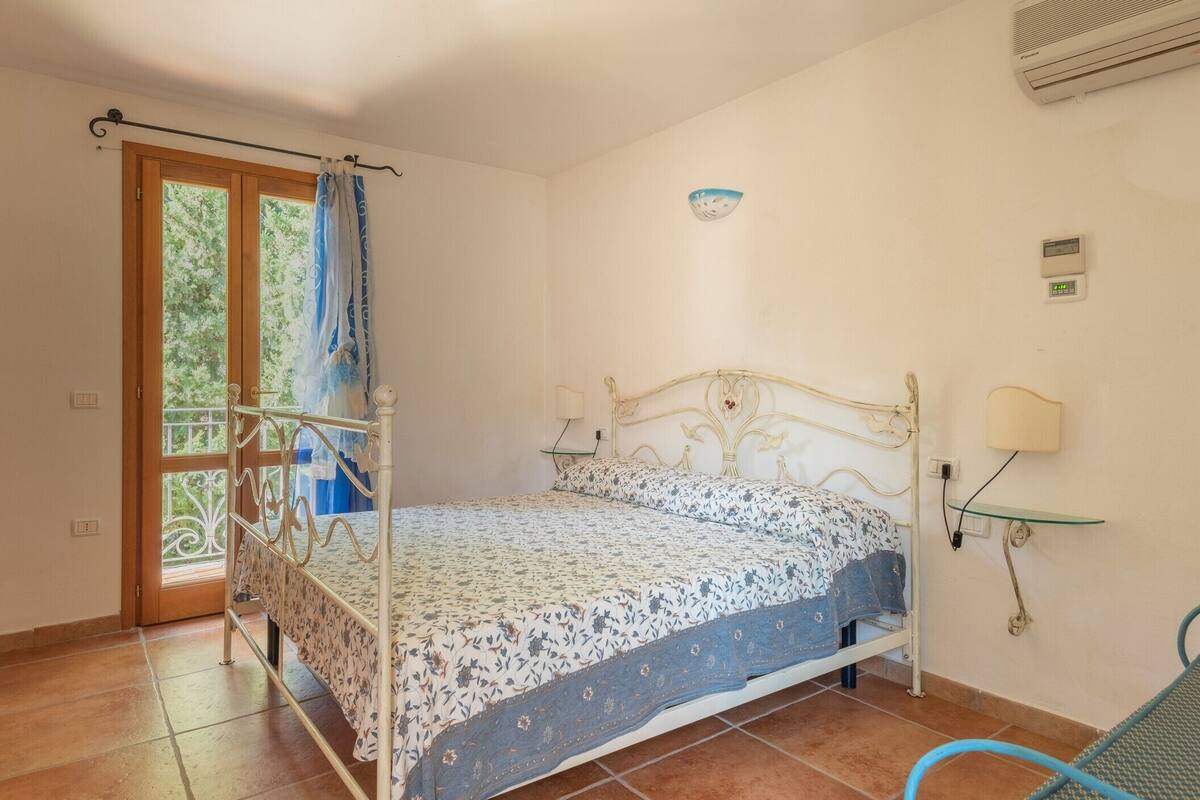 Un dormitorio con estructura de cama metálica, una ventana con cortina, aire acondicionado instalado en la pared y una lámpara de noche.
