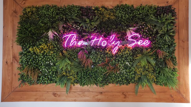 A wall decorated with various plants and a neon sign that reads "The Holy See". - 1 Bedroom Apartment - 1 Bathroom - Large Living Room - San Pietro Station (Roma)