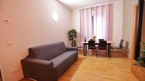 A modern living room with a sofa, a dining table with chairs, a potted plant, and a window with curtains.