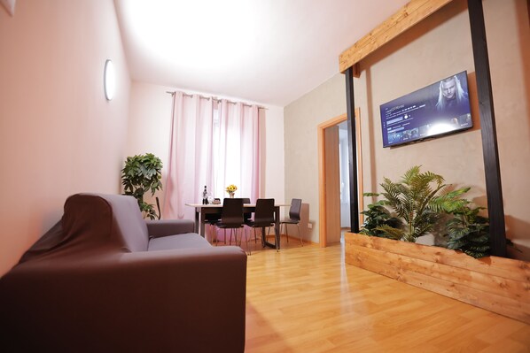 Una sala de estar moderna con un sofá, una mesa de comedor con sillas, un televisor de pantalla plana montado en la pared y piso de madera.