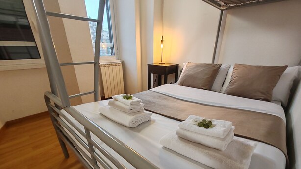 A neatly made bed with two towels, a bedside table with a lamp, and a window with a view of buildings.