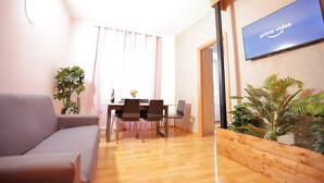 A modern living room with a sofa, dining table, chairs, a TV mounted on the wall, and potted plants.