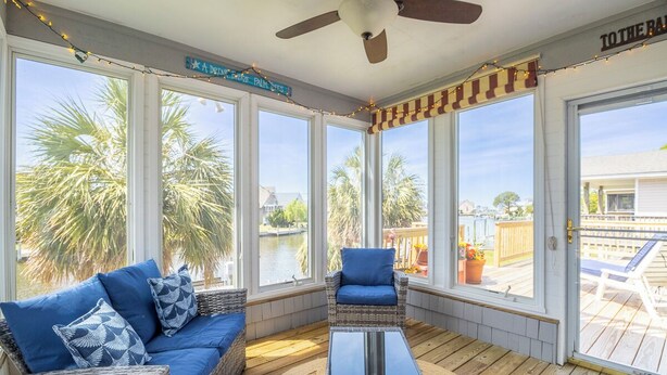 A covered outdoor area with wicker furniture, large windows, and a view of palm trees and a body of water. - Canalfront home w/ wraparound decks and community amenities! (Kill Devil Hills)