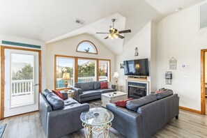 A spacious living room with a high ceiling, a fireplace, a television, and a balcony with a view of houses. - Oceanside home in Duck NC with private mini golf course! (Duck)