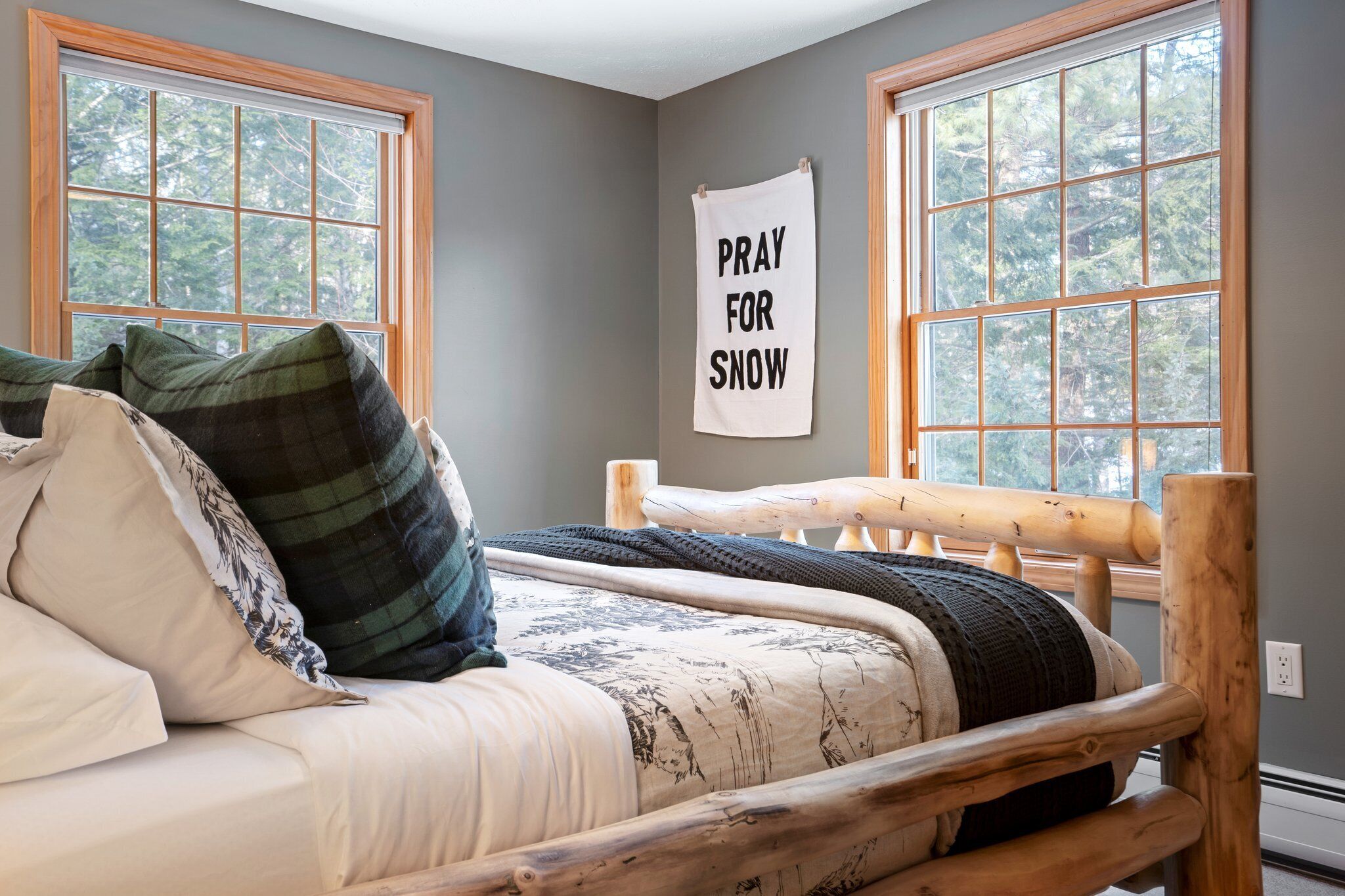 A bedroom with a wooden bed, two windows, and a 'Pray for Snow' sign.