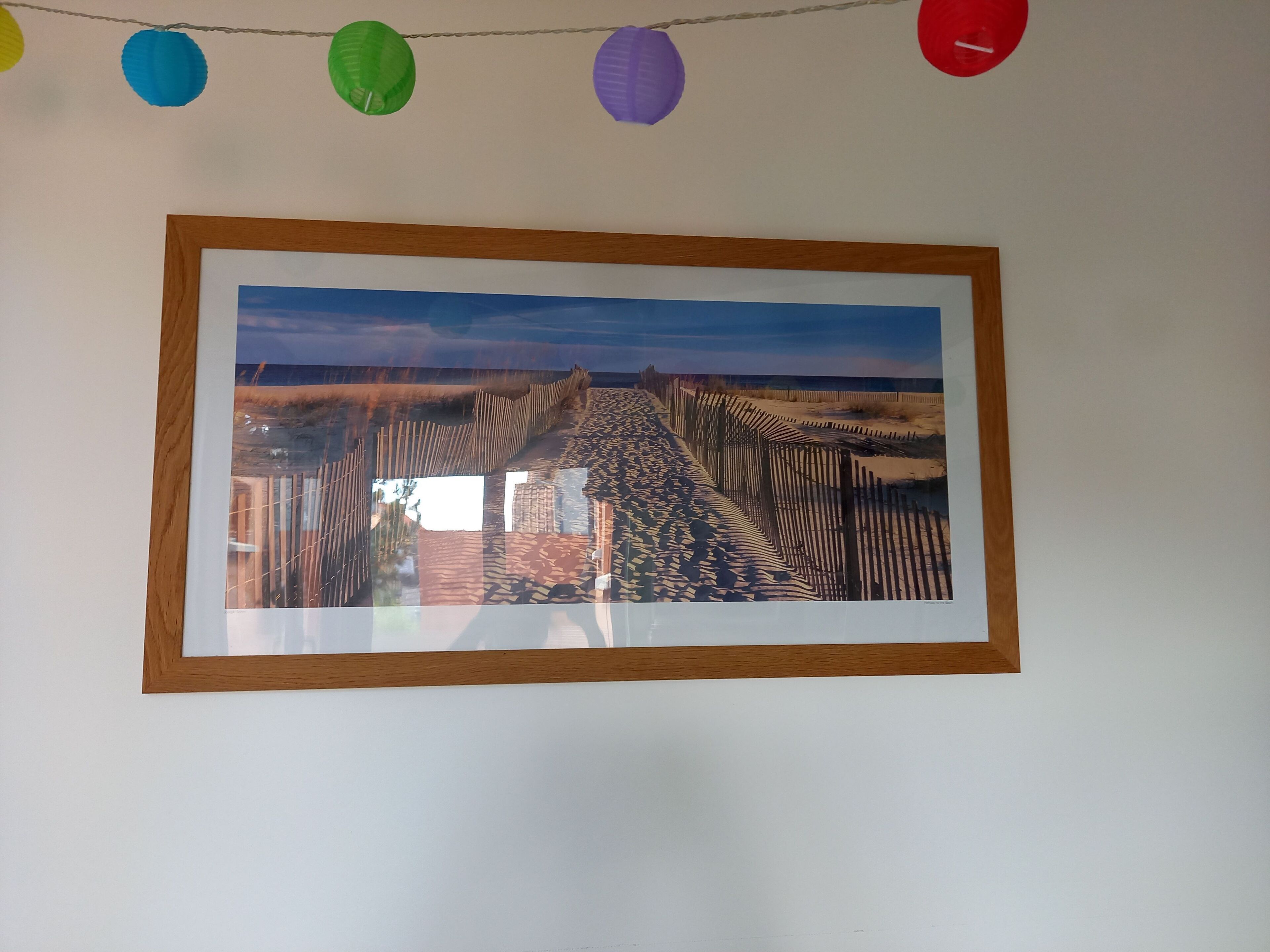 Un cuadro enmarcado de un paisaje costero con una cerca de madera, colgado en una pared blanca.