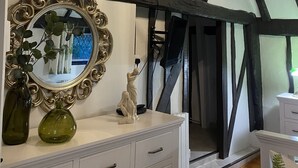 Een kamer met een witte dressoir, een spiegel met gouden lijst, een groene vaas en een beeldje. Er hangt een kroonluchter met meerdere lampen. De kamer heeft zichtbare houten balken in het plafond.