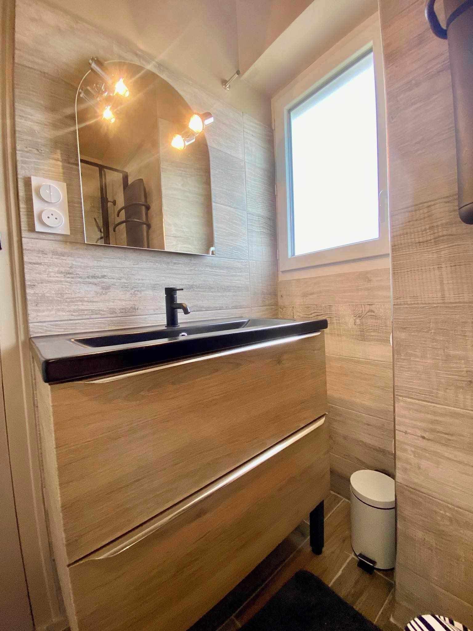 A modern bathroom with a wooden vanity, a black sink, a mirror with lights, and a window.