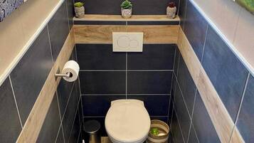 A small bathroom with a toilet, a hand dryer, a trash bin, and a window.