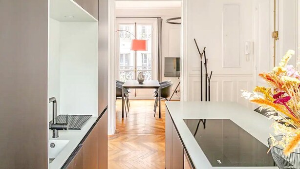 A modern kitchen with a central island, a dining area with a table and chairs, and a view of the street through an open door. - Apartment Thorigny - Two Bedroom Villa, Sleeps 6 (Paris)