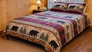 Une chambre à coucher dans un chalet en bois, avec un lit orné d’une courtepointe à motifs d’ours, de grandes fenêtres et un plafond en bois.