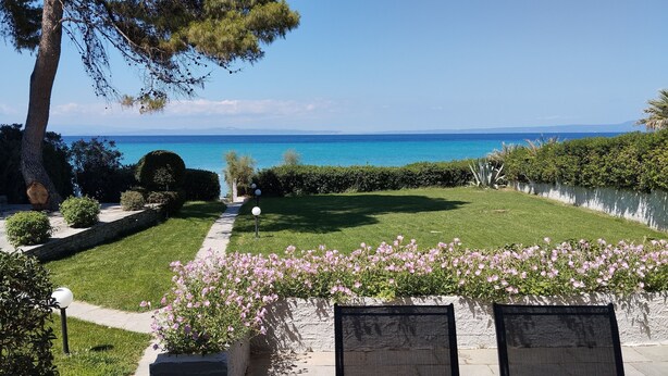 A garden with a pathway leading to a seating area with chairs, a view of the sea, and a hedge with pink flowers.