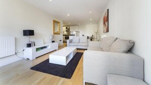 Una sala de estar moderna con un sofá blanco, una mesa de centro blanca y un tapete negro. Se observa una cocina al fondo con gabinetes blancos y una encimera de madera.